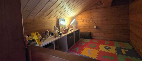 A room with a sloped ceiling, wooden walls, a desk with a lamp, a bed with a colorful mat, and toys.
