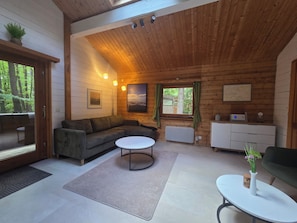 Un intérieur de chalet chaleureux avec un canapé d’angle, une table basse ronde, une petite table blanche, un meuble et une fenêtre avec des rideaux.