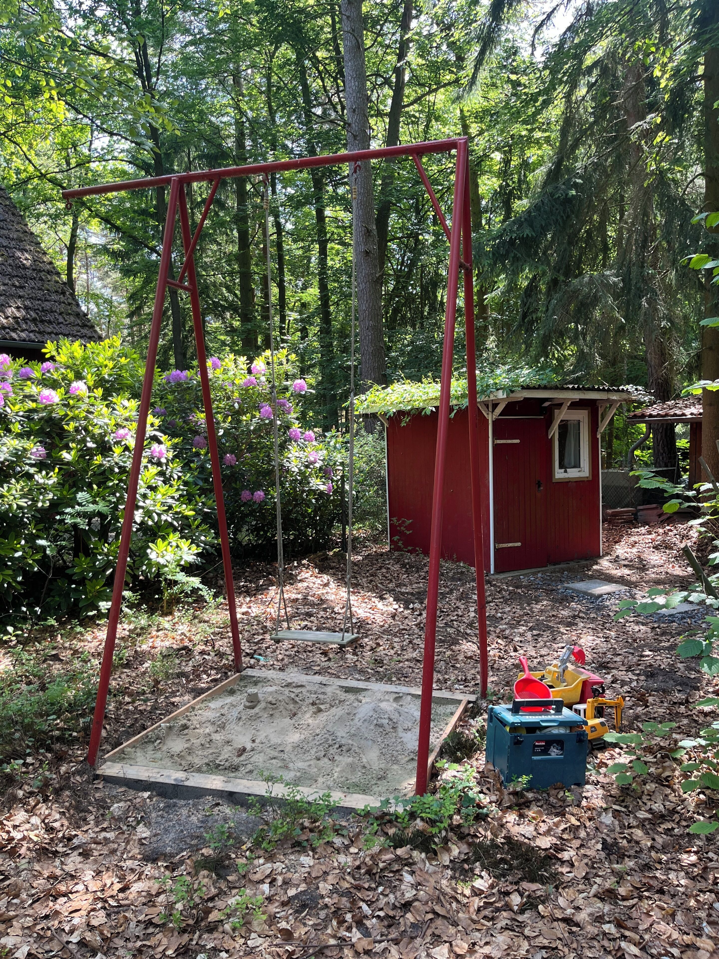 A red play structure in a wooded area with a small shed and a thatched-roof building.