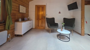 A cozy room with wooden walls, a white dresser, a TV mounted on the wall, two chairs, a small table, and a green curtain. - Idyllic wooden house on the Steinhuder Meer (Neustadt am Rübenberge)