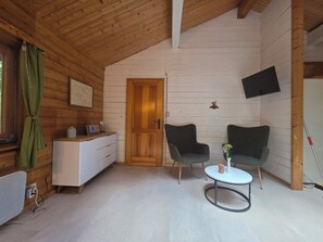 Une pièce chaleureuse avec des murs en bois, une commode blanche, un téléviseur fixé au mur, deux chaises, une petite table et un rideau vert.