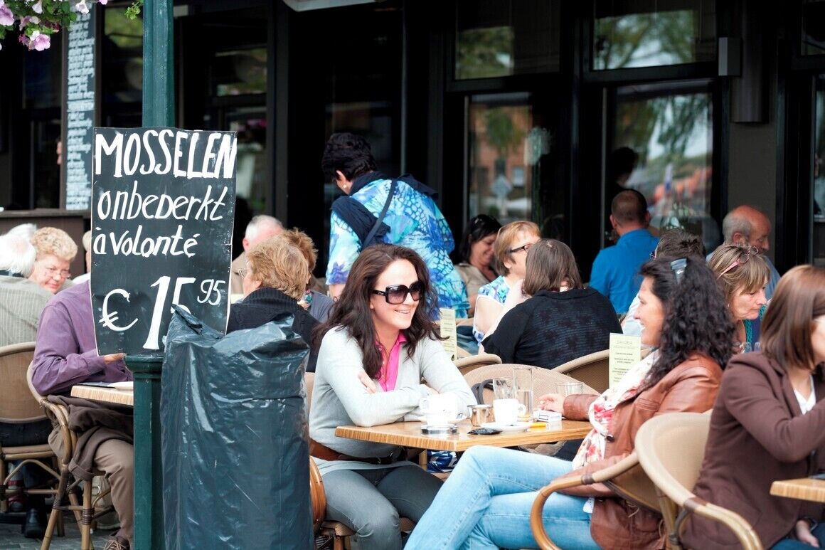 An outdoor café with people seated at tables, a sign with text, and a trash bin.