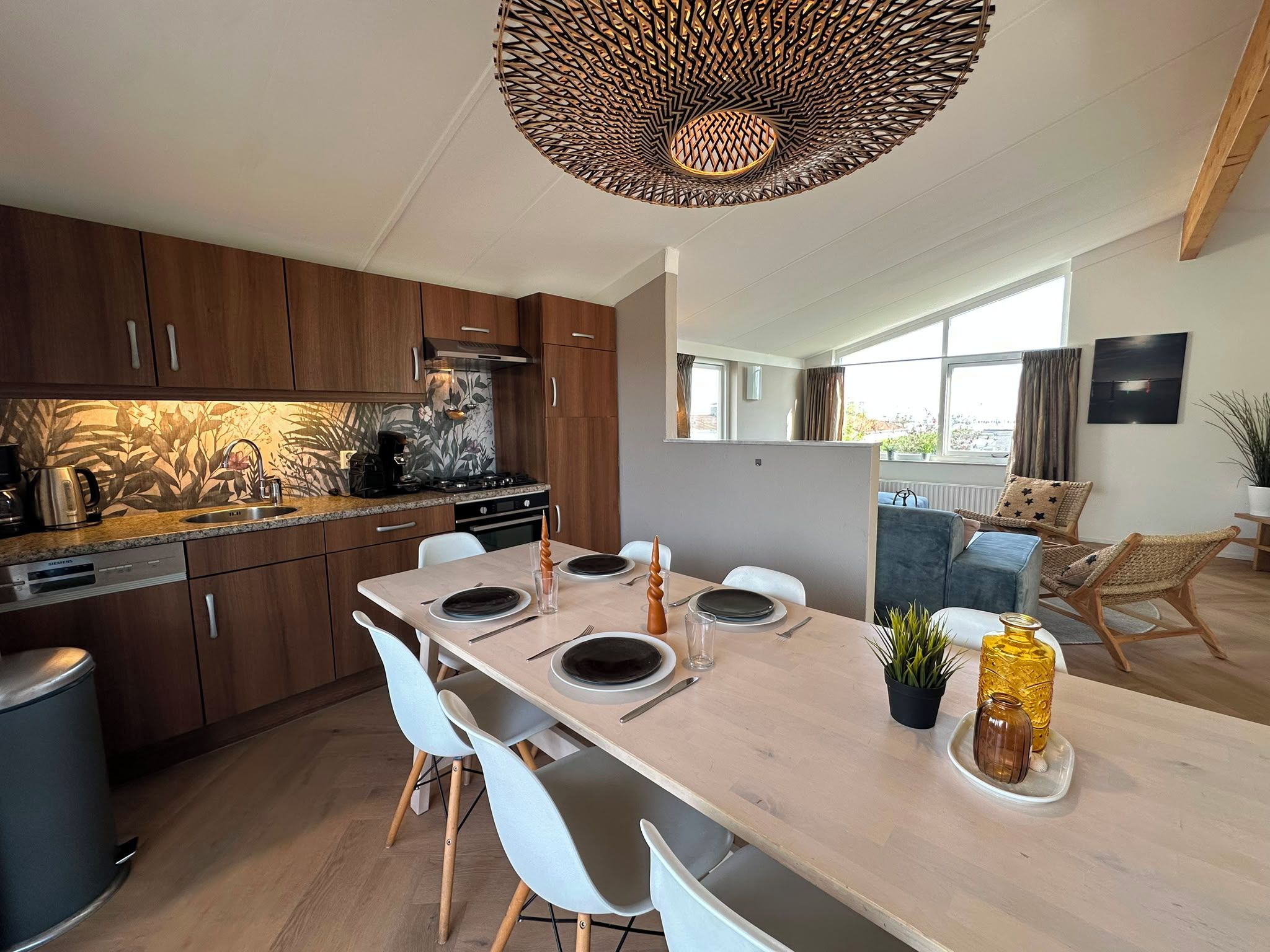 A modern kitchen with wooden cabinets, a dining area with a long table, and a view of the outdoors through a large window.