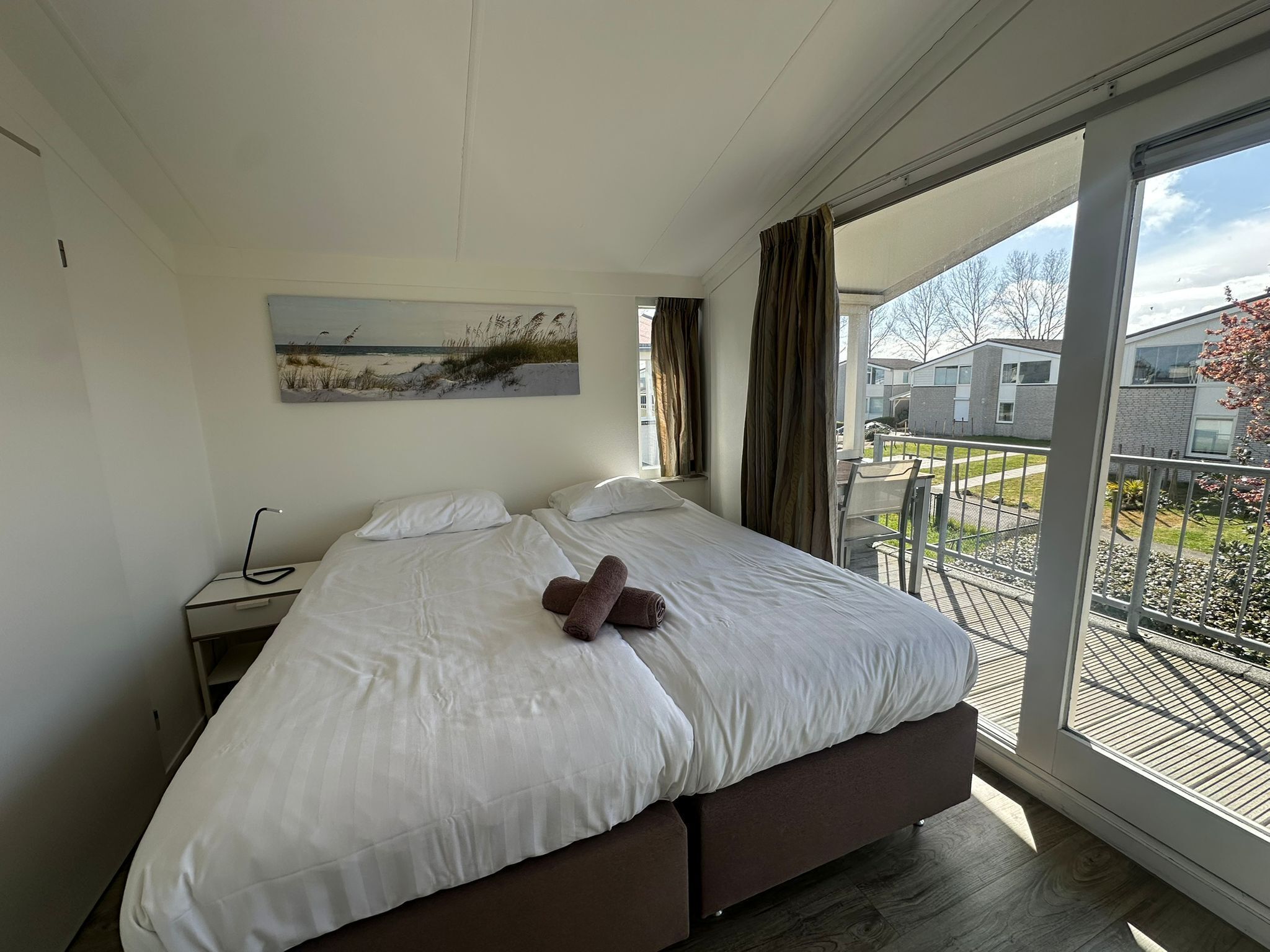 A bedroom with a large bed, a nightstand, a painting of a beach, and a view of a building through a glass door.
