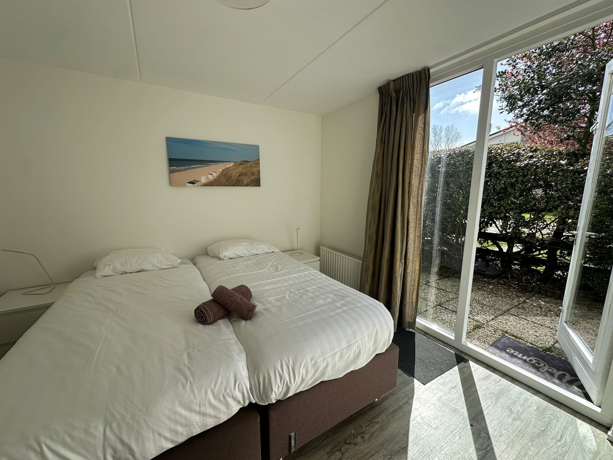 A bedroom with a bed, a painting of a beach, and a view of a garden through a glass door.
