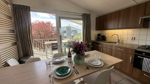Uma área de refeições com mesa posta para quatro, uma cozinha com armários de madeira e vista para uma varanda com folhagem de outono.