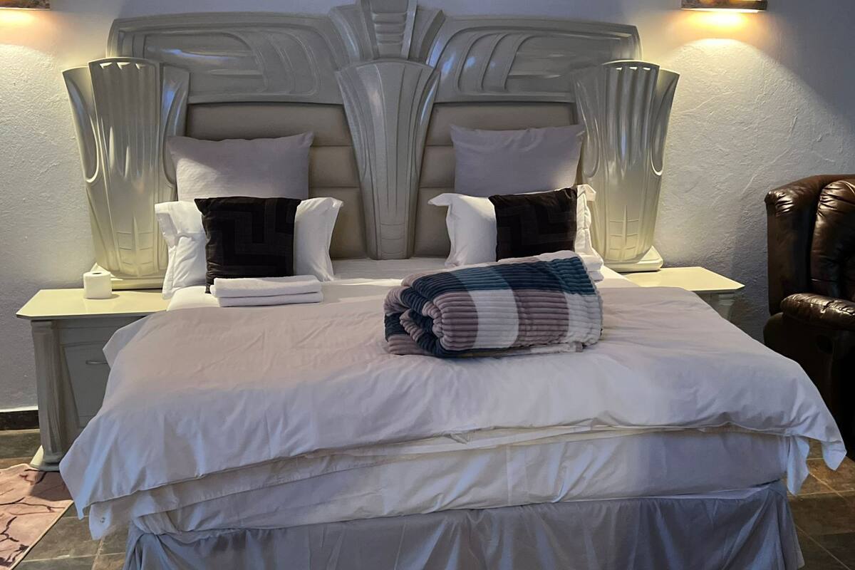 Una cama bien hecha con sábanas blancas, un edredón doblado y dos almohadas. Hay dos mesitas de noche con lámparas y una butaca de cuero marrón.
