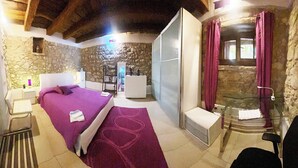 A bedroom with a stone wall, a bed with a purple bedspread, a small bedside table, and a glass-enclosed shower area.