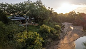 A house with a covered patio, surrounded by dense greenery and a river. - Nomads Den - Safari Villa North - Private heated pool and riverbed views (Hoedspruit)