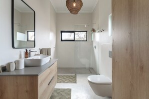 A modern bathroom with a large mirror, a freestanding sink, a toilet, and a glass-enclosed shower. - Nomads Den - Safari Villa North - Private heated pool and riverbed views (Hoedspruit)