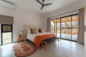 A bedroom with a bed, a bench, a nightstand, a floor lamp, a ceiling fan, and a large window with curtains. - Nomads Den - Safari Villa North - Private heated pool and riverbed views (Hoedspruit)