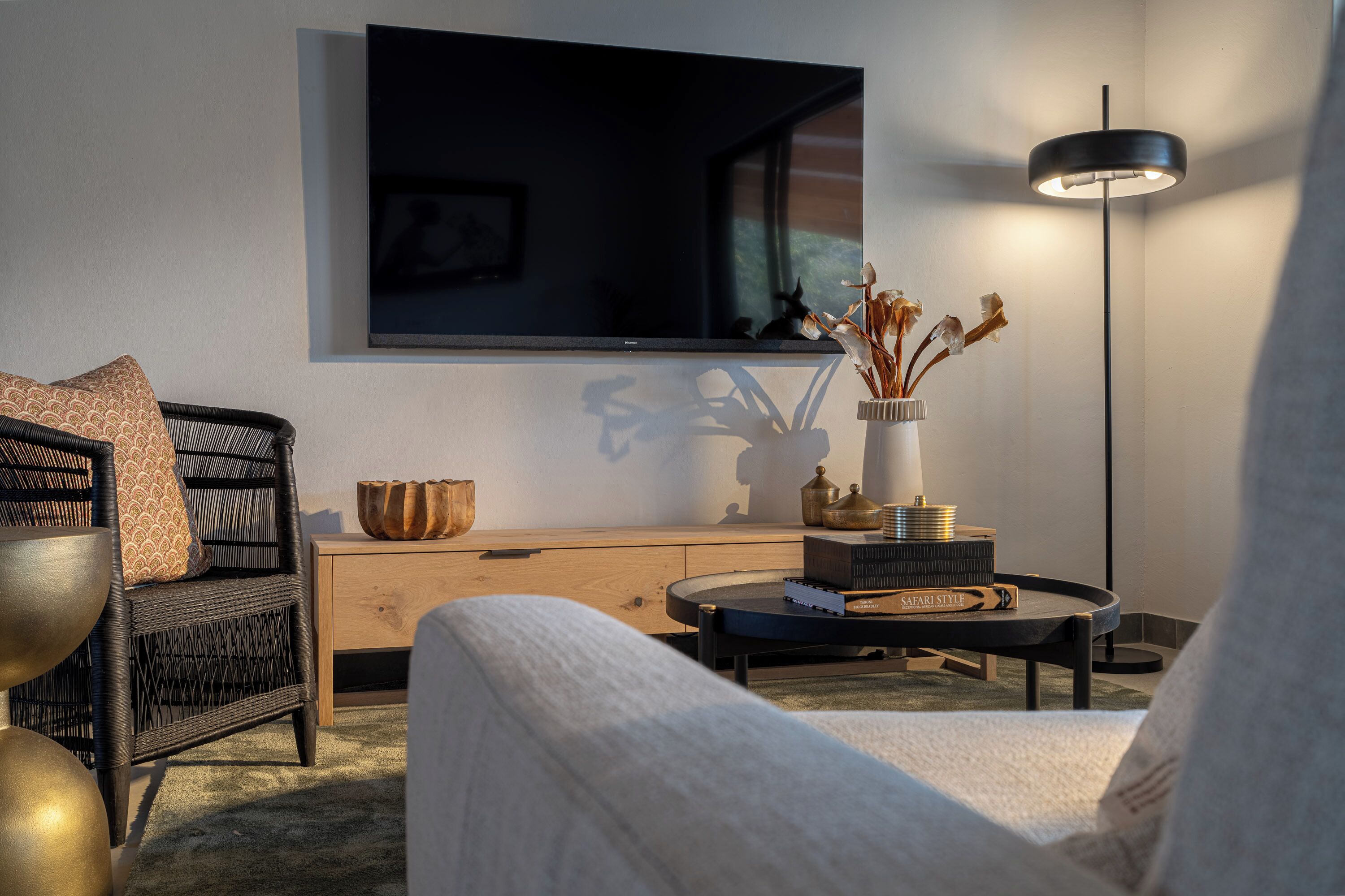 Un salon moderne comprenant un canapé, un fauteuil, une table basse, un téléviseur, un lampadaire et un vase avec des fleurs séchées.