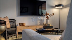 A modern living room with a sofa, a chair, a coffee table, a TV, a floor lamp, and a vase with dried flowers. - Nomads Den - Safari Villa North - Private heated pool and riverbed views (Hoedspruit)