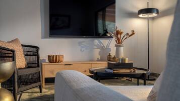 Un salon moderne comprenant un canapé, un fauteuil, une table basse, un téléviseur, un lampadaire et un vase avec des fleurs séchées.