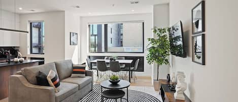 A modern living room with a sofa, a dining table, and chairs, a kitchen area, and a large window with a city view.