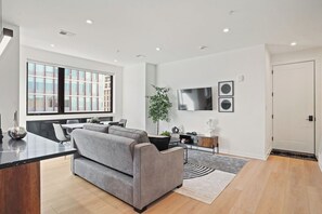 Una sala moderna con un sofá gris, una zona de comedor con mesa y sillas, un ventanal grande y un televisor de pantalla plana montado en la pared.