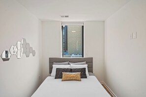 A bedroom with a bed, a window, a wall-mounted mirror, and a ceiling vent.