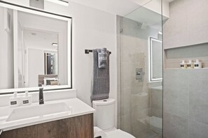 Un baño moderno con ducha envuelta en vidrio, un lavamanos blanco con tocador de madera y un espejo grande.