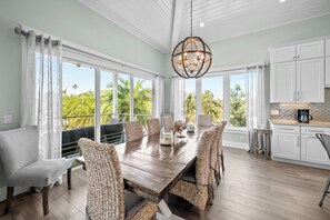A dining area with a wooden table and wicker chairs, large windows with a view of palm trees, and a kitchen with white cabinets. - Casa Azul by Beach Boutique Rentals (Anna Maria)