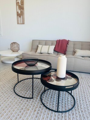 Ein Wohnzimmer mit einem grauen Sofa, zwei runden Tischen mit schwarzen Rahmen, einer weißen Vase und einem gewebten Teppich.