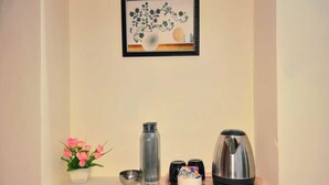 A wall-mounted picture frame with a floral design, a small potted plant with pink flowers, a glass bottle, a metal cup, and a stainless steel kettle on a shelf. - The Green Homez (Bengaluru)