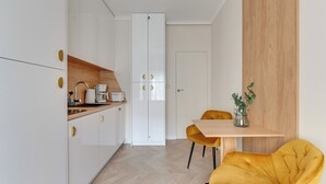 A compact kitchen with white cabinets, a wooden countertop, a coffee maker, and a dining area with a yellow chair and table. - Sea & Monte City Center Apartments Sopot (Sopot)