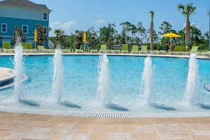 Una piscina con múltiples chorros de agua, sillas reclinables verdes y palmeras.