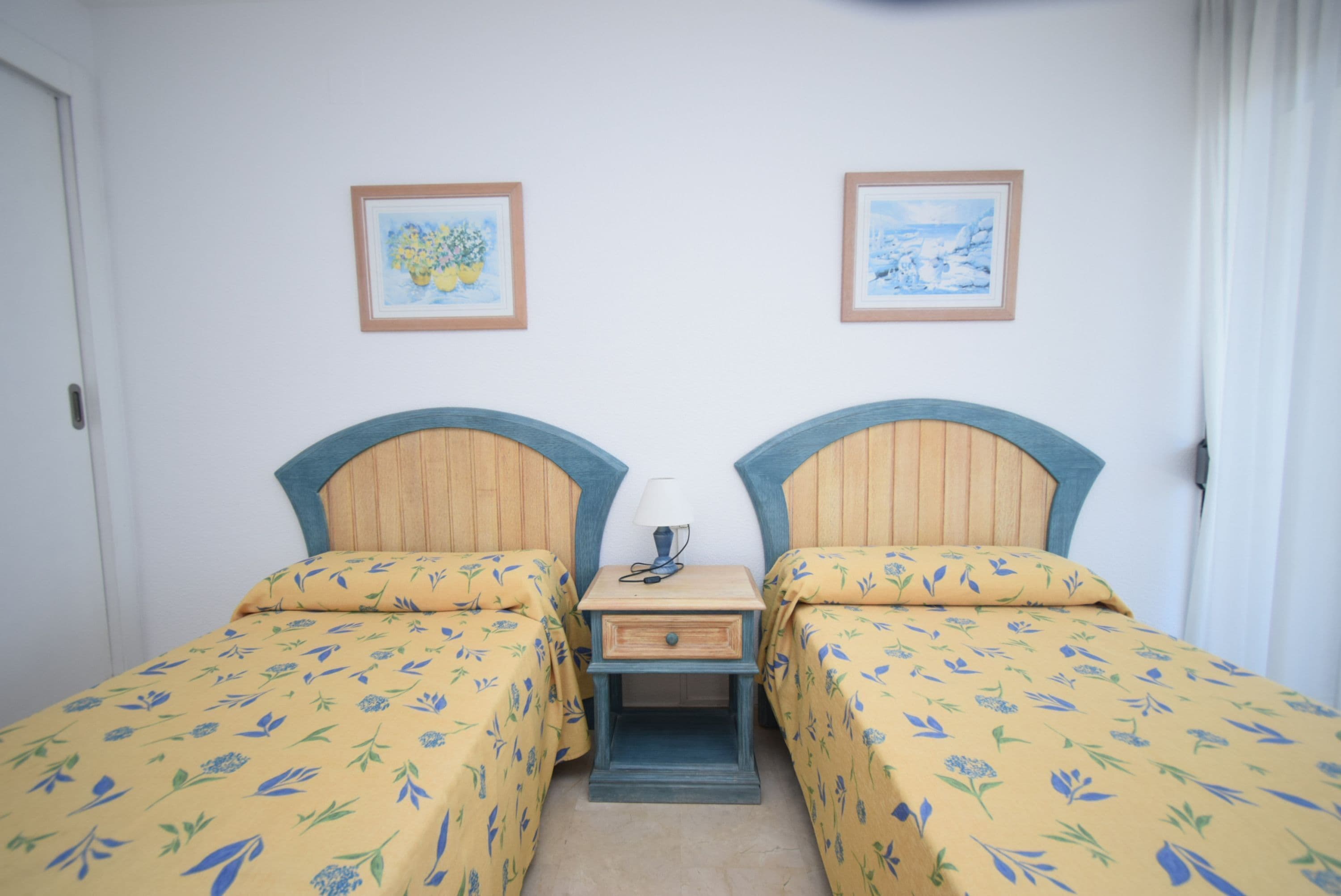 Een kamer met twee bedden, elk met een houten hoofdbord, een nachtkastje met een lamp en ingelijste foto’s aan de muur.