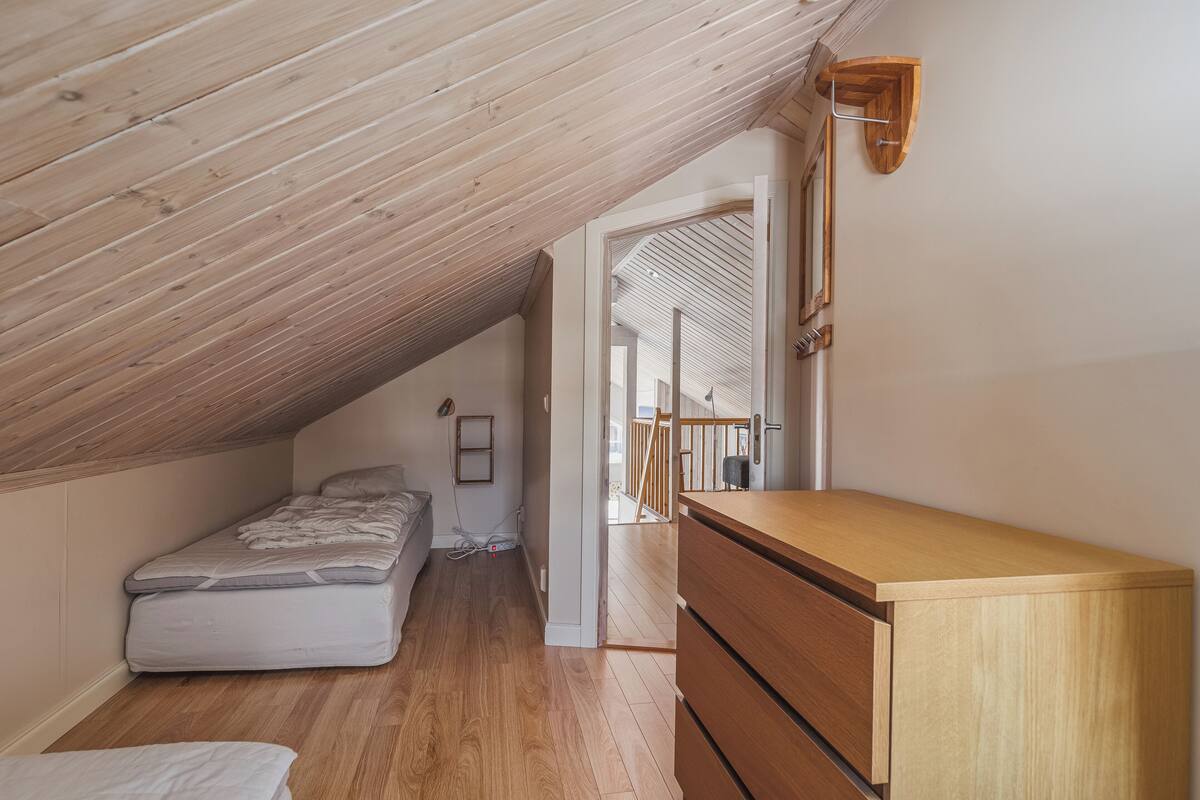Een kleine kamer met een schuine kap, een houten vloer, een bed, een dressoir en een houten wandlamp.