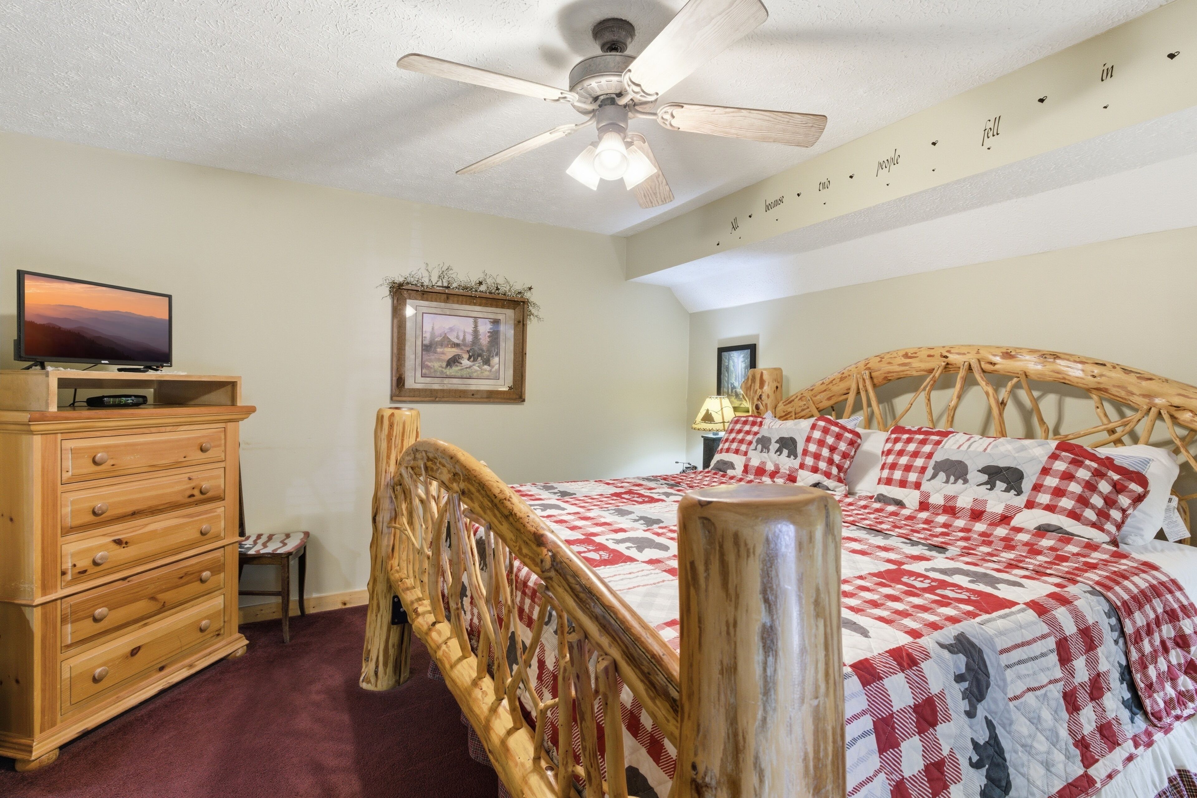 A bedroom with a wooden bed, a dresser, a television, a ceiling fan, and a wall with a decorative phrase.