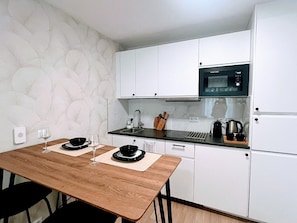 Une cuisine compacte avec des meubles blancs, un four à micro-ondes, un évier et une table à manger avec deux assiettes et des verres à vin.