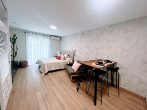 Une pièce de taille modeste comprenant un lit, une table à manger avec des chaises et un mur orné d’un motif texturé.