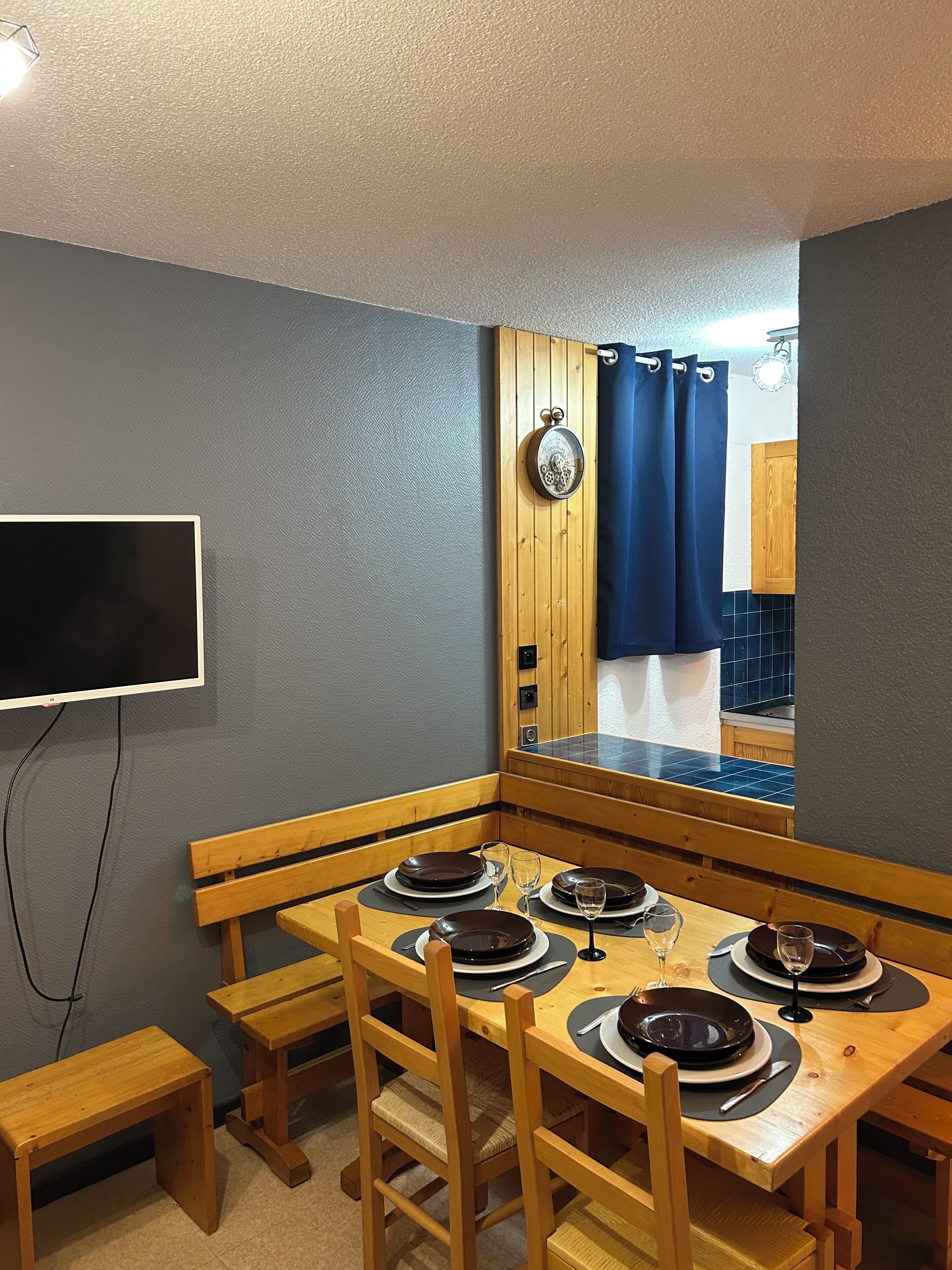 A dining area with a wooden table set for four, a television mounted on the wall, and a wooden cabinet in the background.