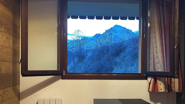 Ein Schlafzimmer mit großem Fenster, einem Bett mit rot-weiß gestreifter Bettdecke, einem Heizkörper und einem schwarzen Lautsprecher.