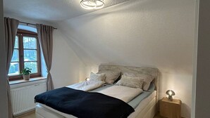 Makuuhuoneessa on sänky, yöpöytä, ikkuna, jossa on verhot, ja kattovalaisin.