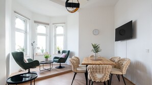 A modern living room with a wooden dining table, green and beige chairs, a round coffee table, and a TV mounted on the wall. - Faraway Homes Studio Klosterneuburg #5 (Klosterneuburg)
