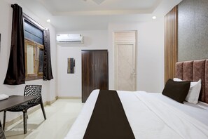 A hotel room with a bed, a desk, a chair, a wardrobe, and an air conditioning unit.