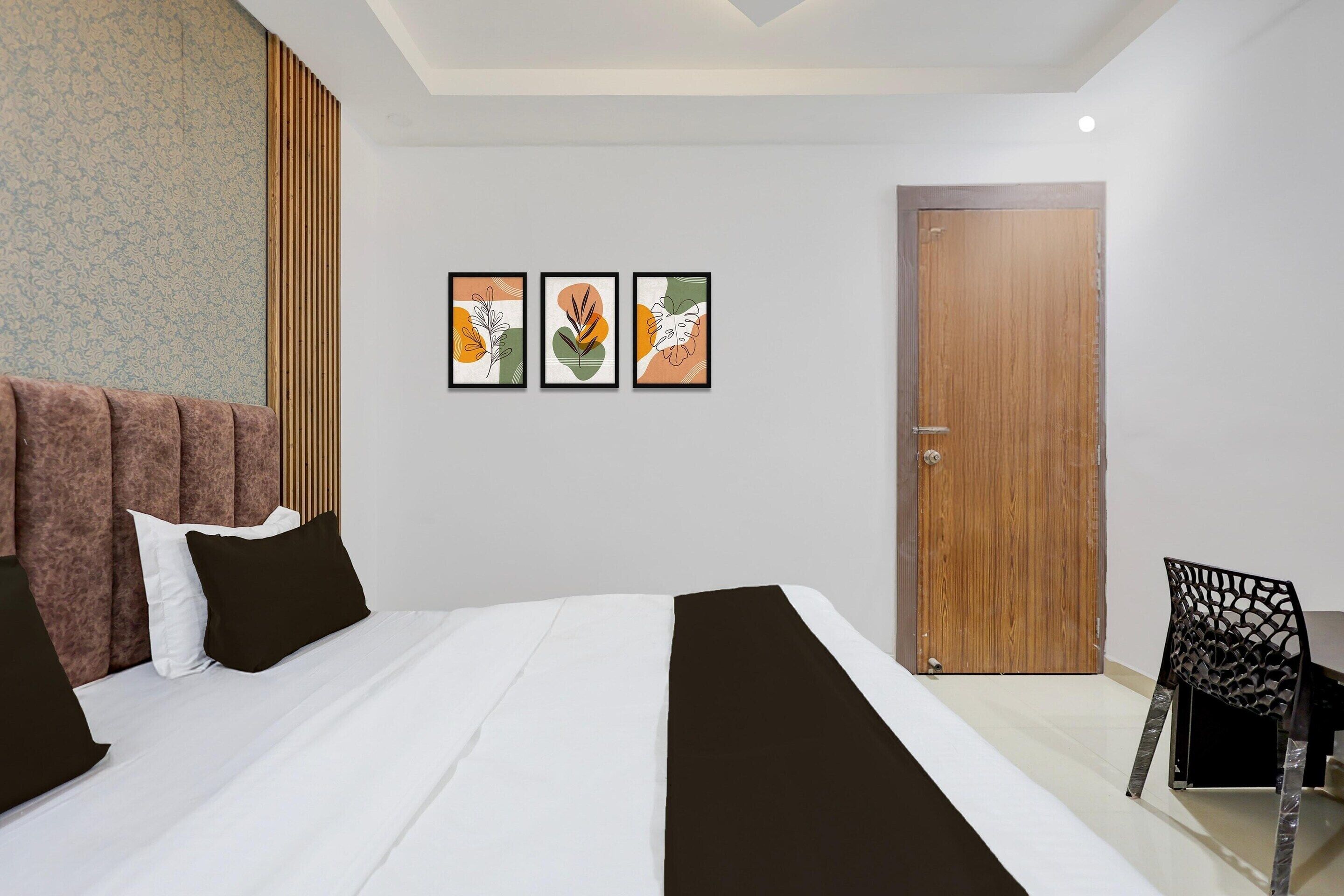 A hotel room with a bed, a wooden door, a chair, and three framed artworks on the wall.