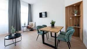 Un salon moderne avec une table à manger en bois, des chaises recouvertes d’un tissu vert, un fauteuil blanc, une petite table d’appoint noire et une bibliothèque en bois.