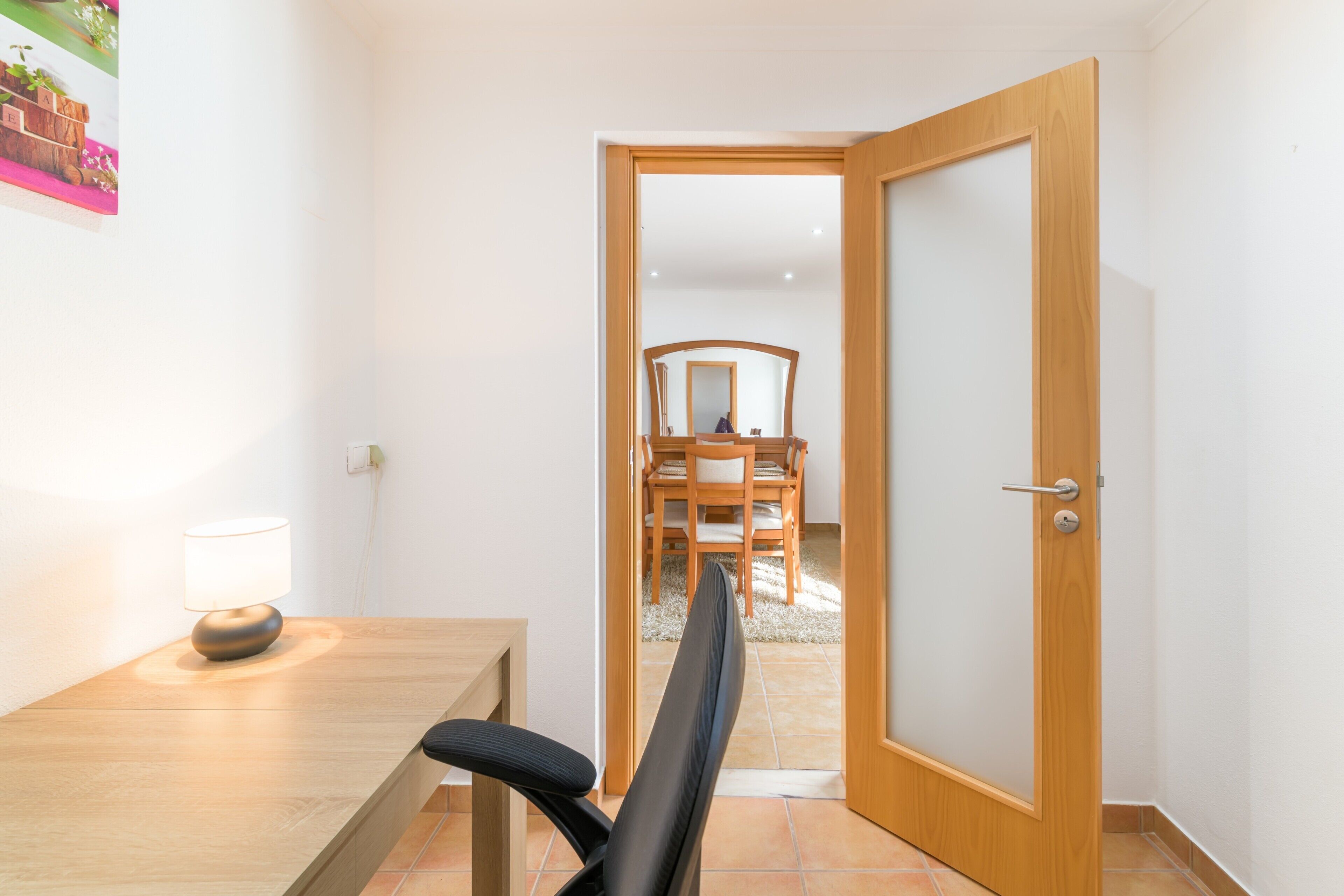 Una habitación con un escritorio de madera, una silla, una lámpara y una puerta que da a un comedor con sillas y una mesa.