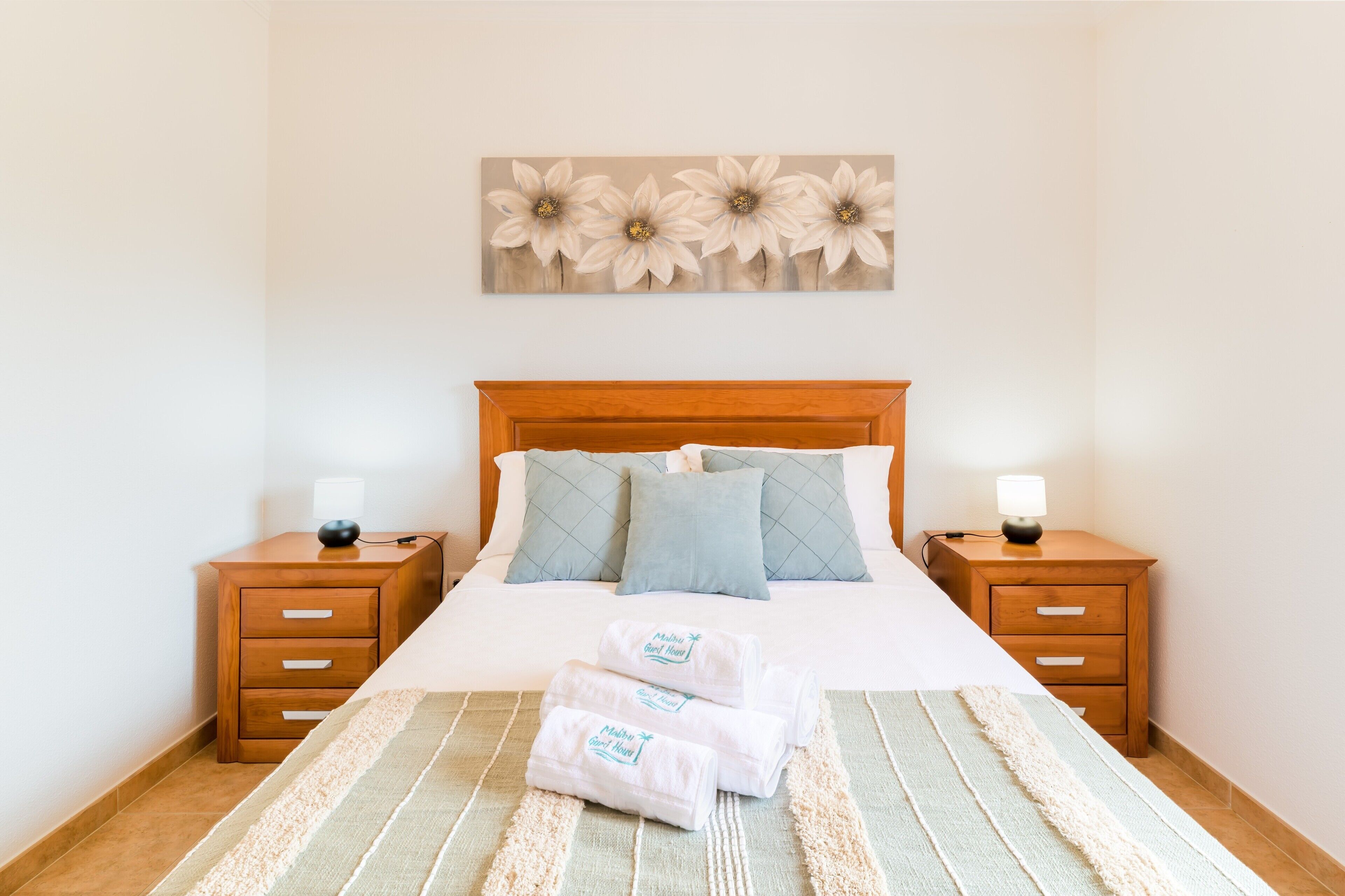 Un letto rifatto con una coperta a righe, due comodini con lampade, un quadro di fiori appeso al muro e un piccolo cassetto.