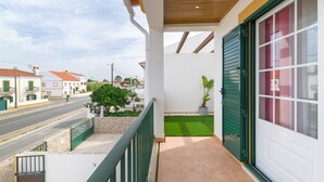 Un balcone con vista su una strada, una ringhiera verde, una pianta in vaso e una porta con persiane verdi.