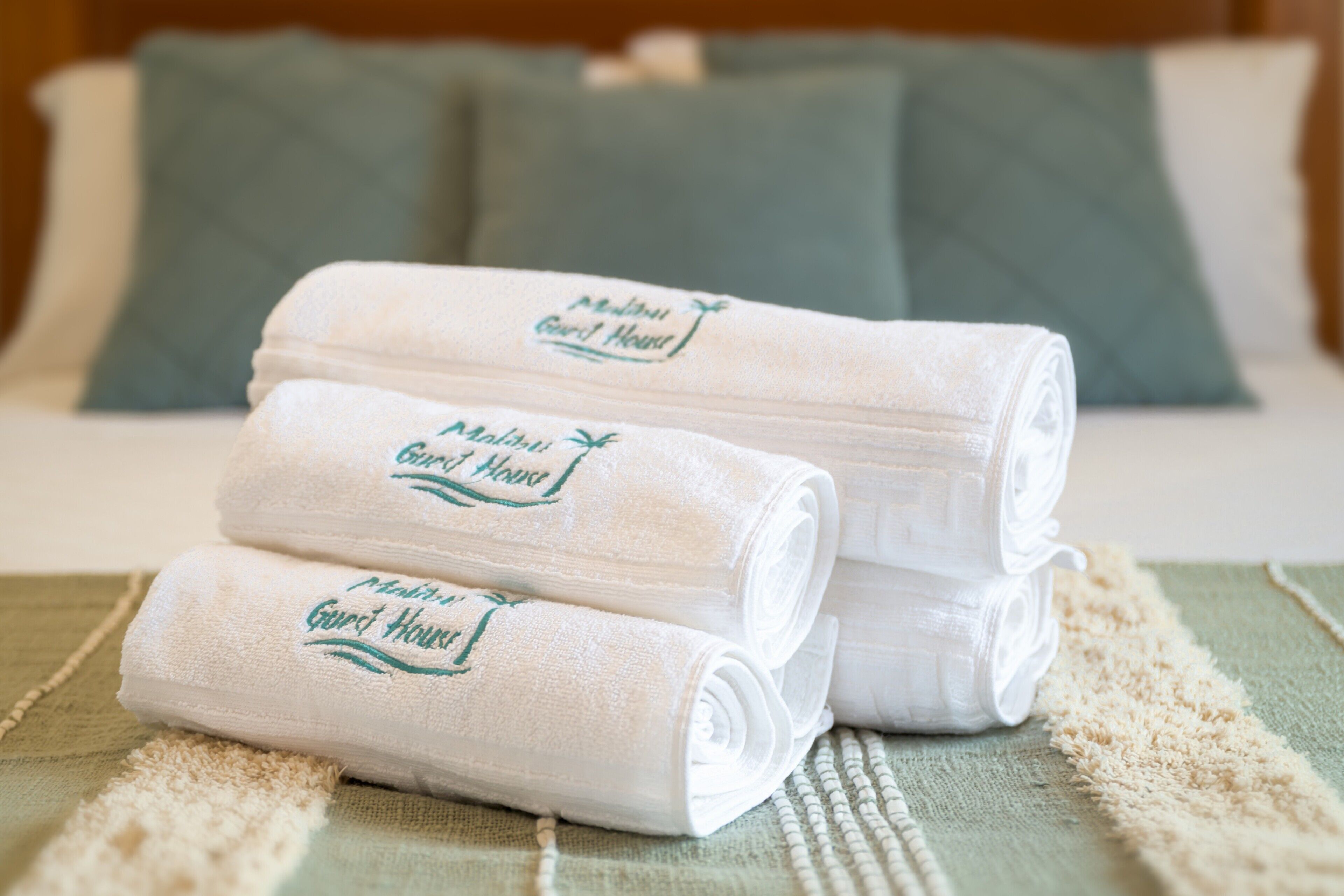 Snyggt hopvikta handdukar med logotypen för ”Malibu Guest House” ligger på en säng med grönt sänglinne och kuddar.