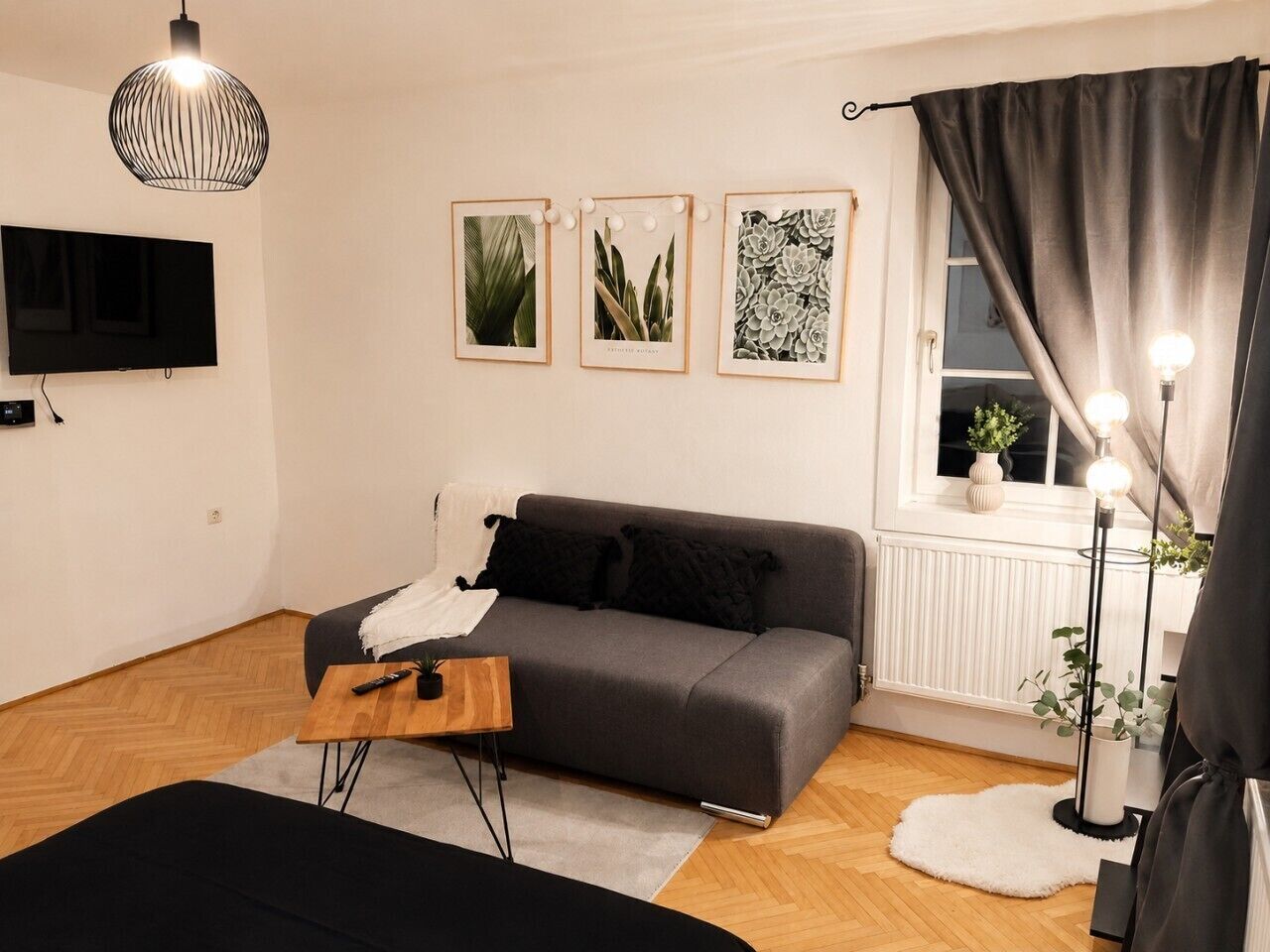 A modern living room with a grey sofa, a wooden coffee table, a TV mounted on the wall, and framed botanical prints.