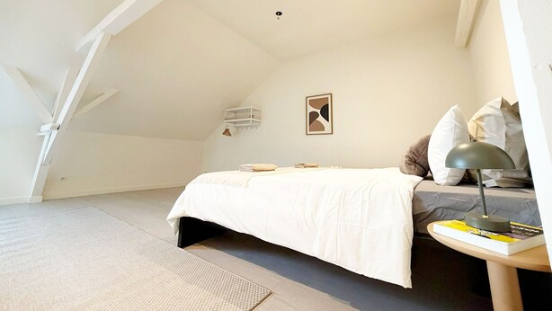 Een slaapkamer met een bed, nachtkastje met lamp, klein schapje en een wandlamp.
