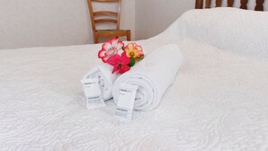 En pænt redt seng med sammenrullet hvide håndklæder og blomsterarrangement, en træstol og en ensfarvet væg.