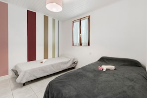Una stanza con due letti, uno con copriletto grigio e l'altro con copriletto grigio scuro, una finestra con tende bianche e un soffitto bianco con travi in legno.
