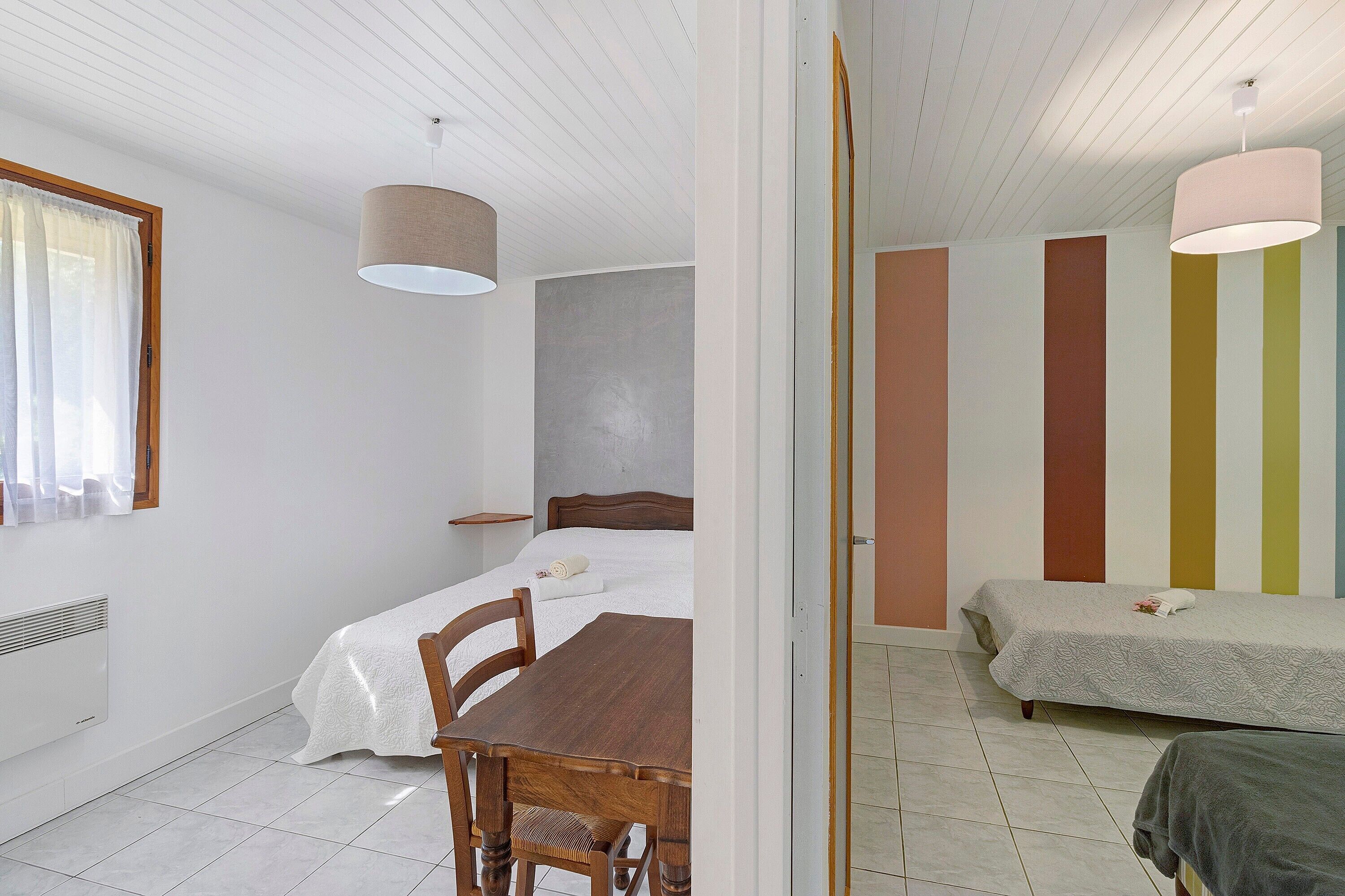 A room with a bed, a table, and a chair. There is a window with curtains, a ceiling fan, and a radiator.