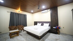 A bedroom with a bed, two chairs, a small table, a nightstand, a vase with flowers, and an air conditioning unit. - Aarna Coastal Resort (Bhatkal)
