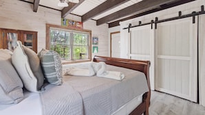 Un dormitorio con cama de madera, ventana con vista a follaje, puerta corrediza y ventilador de techo.